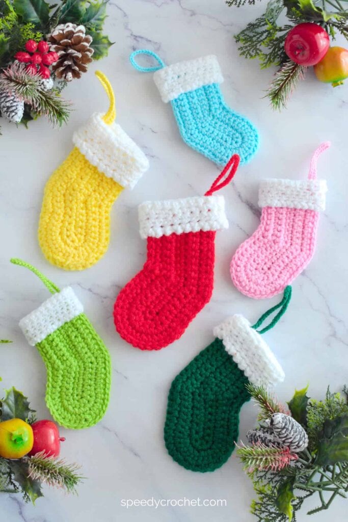 Six small crocheted Christmas stockings in assorted colors with white cuffs, arranged on marble surface with pinecones, holly, berries, and festive decorations.