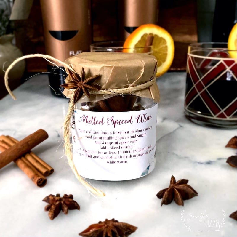 Jar of mulled spice mix wrapped in brown paper with twine and star anise, placed on marble surface with cinnamon sticks, star anise, and glass of red wine garnished with orange slice, copper containers in background.