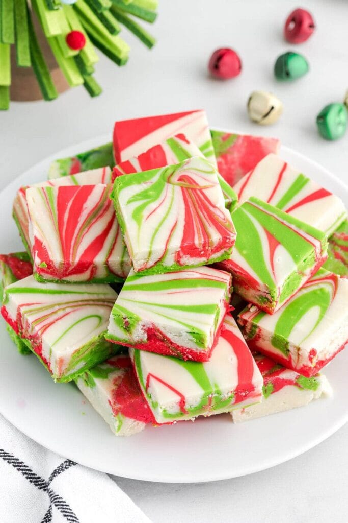Stacked squares of red, green, and white swirled fudge on white plate, surrounded by jingle bells and Christmas tree decorations.