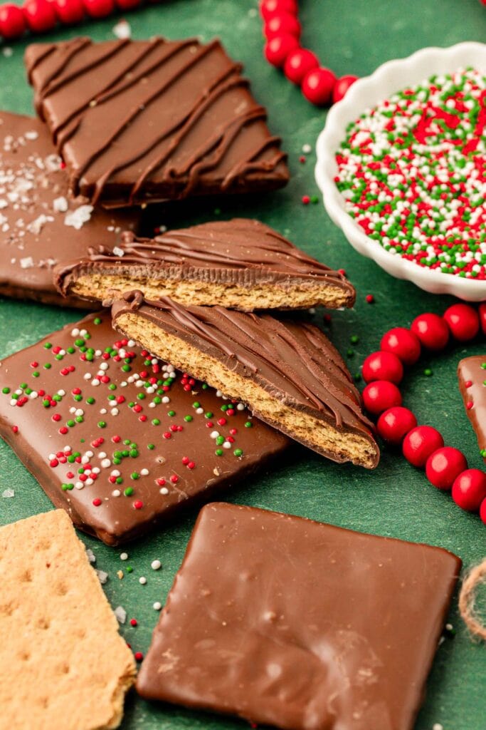 Chocolate-covered graham crackers decorated with sprinkles, drizzle, and sea salt, arranged on green surface with bowl of sprinkles and red bead garland.