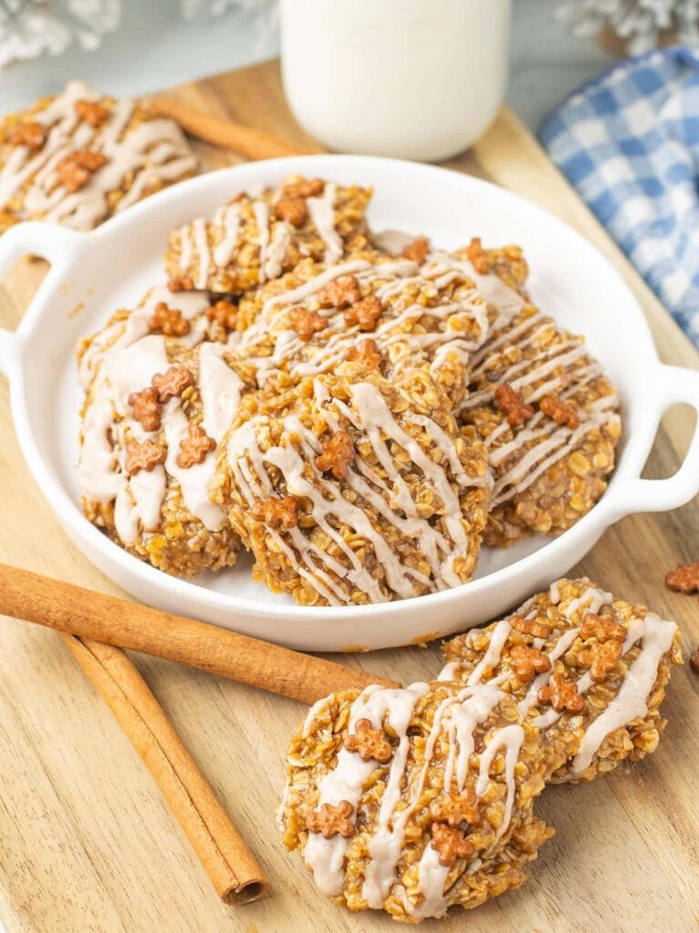 Plate of oatmeal cookies drizzled with icing and topped with flower-shaped sprinkles, beside cinnamon sticks on wooden surface with milk and checkered cloth in background.