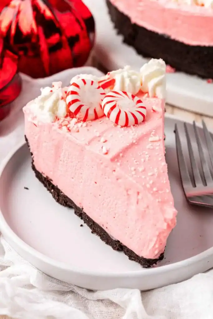 Slice of pink peppermint pie with chocolate crust, whipped cream, striped candies, and crushed peppermint, served on white plate with fork beside festive ornaments and whole pie in background.
