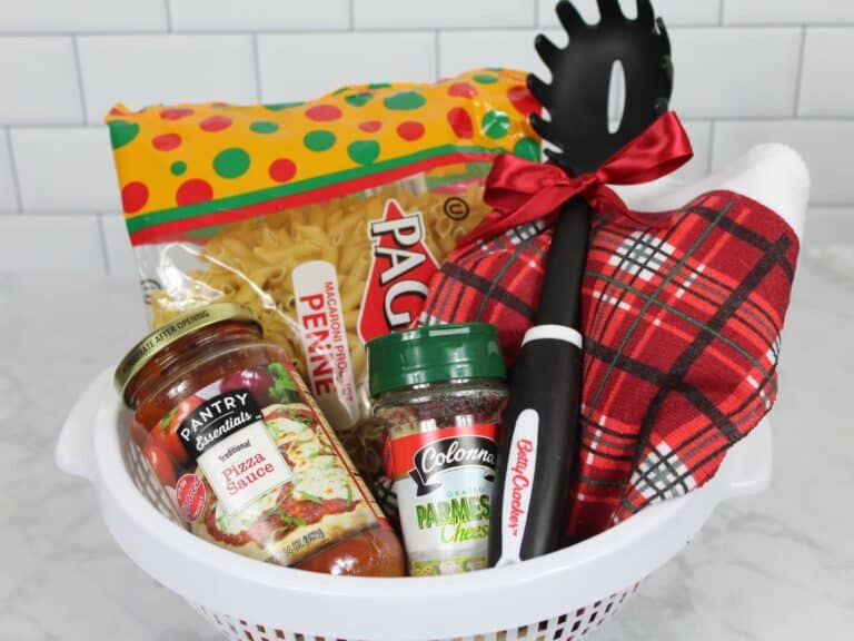 White colander gift basket with pasta, pizza sauce, Parmesan, red plaid towel, and spoon tied with bow, arranged on marble countertop.