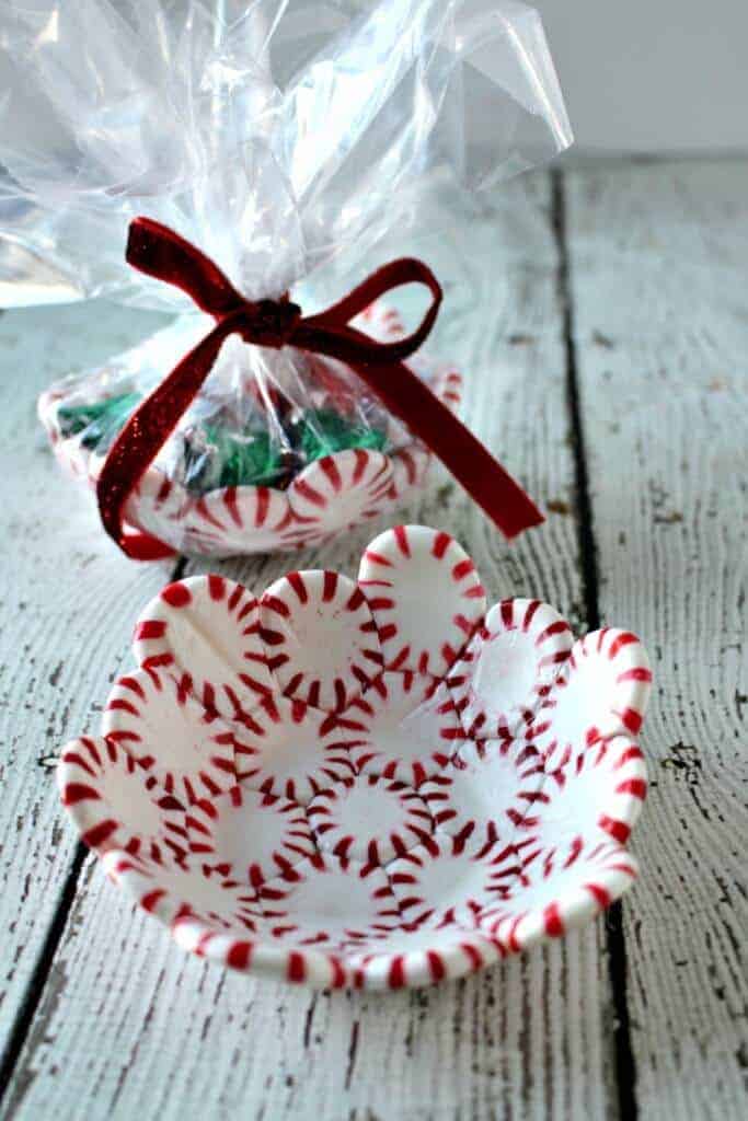 Decorative bowl made from melted peppermint candies with scalloped edges, displayed on rustic white surface beside wrapped version tied with red ribbon and filled with green sweets.
