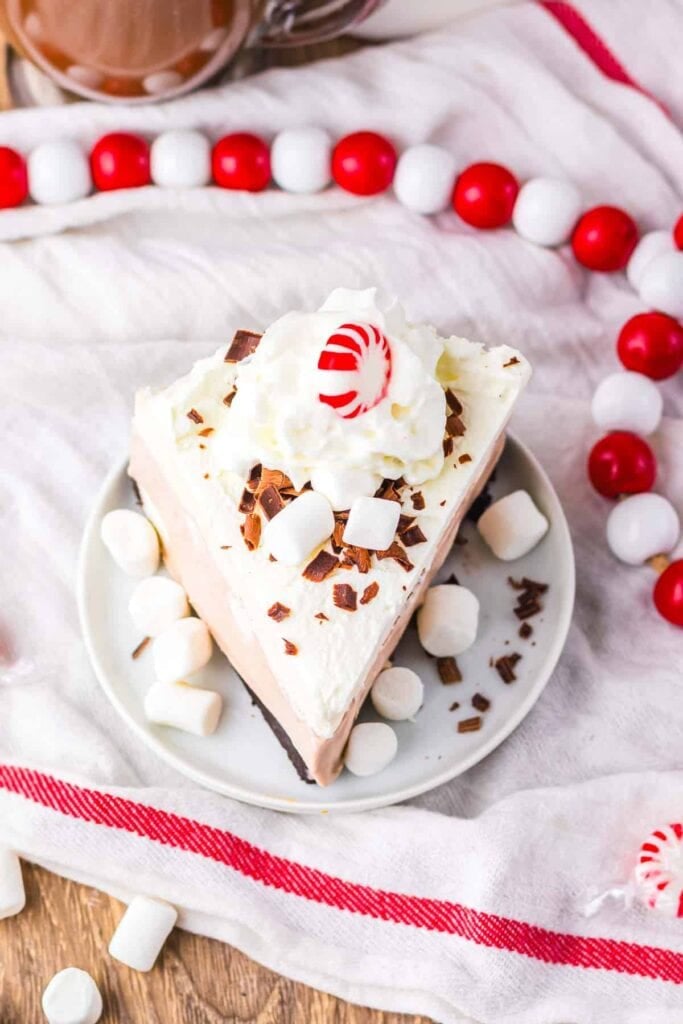 Slice of cream pie topped with whipped cream, chocolate shavings, marshmallows, and peppermint candy, served on white plate with red-striped cloth, scattered marshmallows, and hot chocolate in background.
