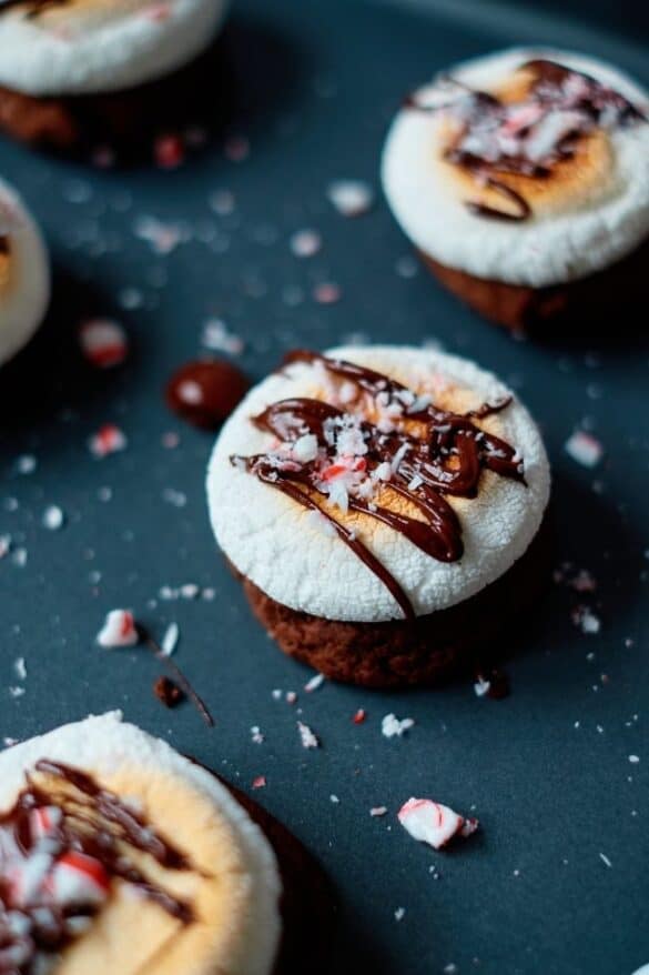 Chocolate cupcakes topped with toasted marshmallows drizzled in chocolate and sprinkled with crushed peppermint, arranged on dark surface with scattered candy pieces.