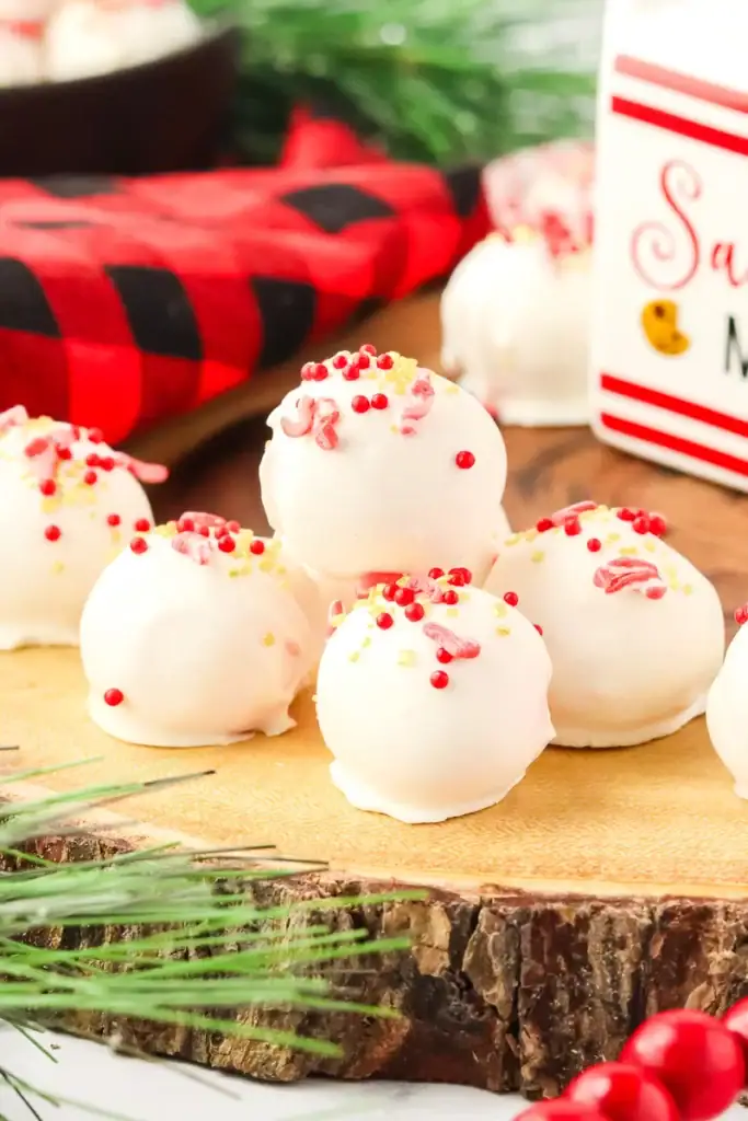 Wooden board of white chocolate truffles with red and yellow sprinkles, surrounded by pine branches, red berries, and Christmas décor.