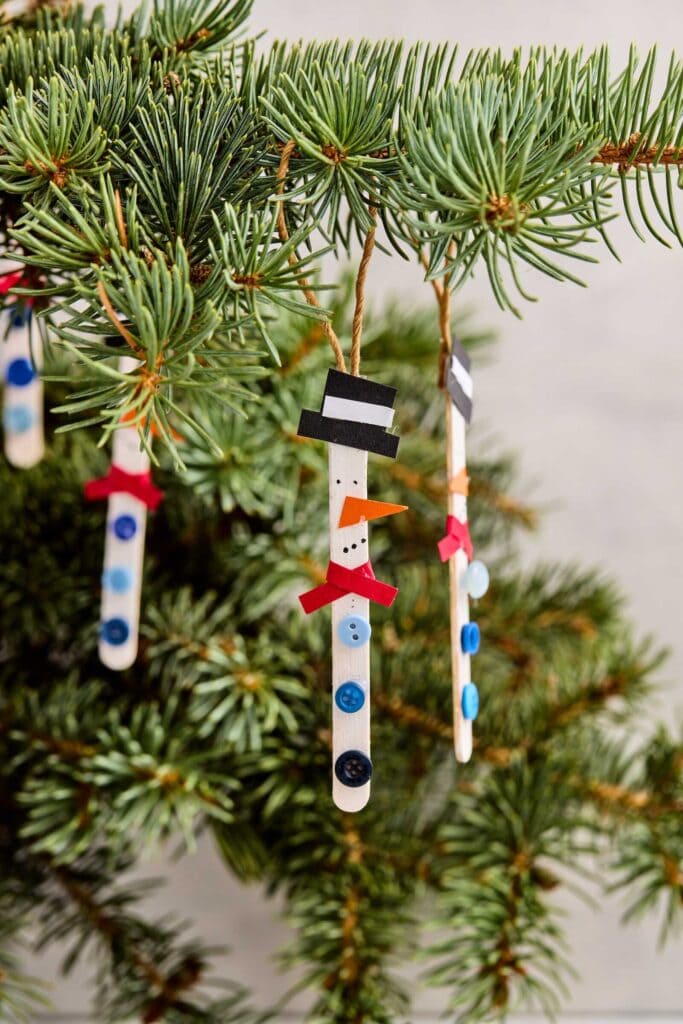 Handmade snowman ornaments from white popsicle sticks with hats, noses, buttons, and ribbon bows, hanging on pine branch with twine.