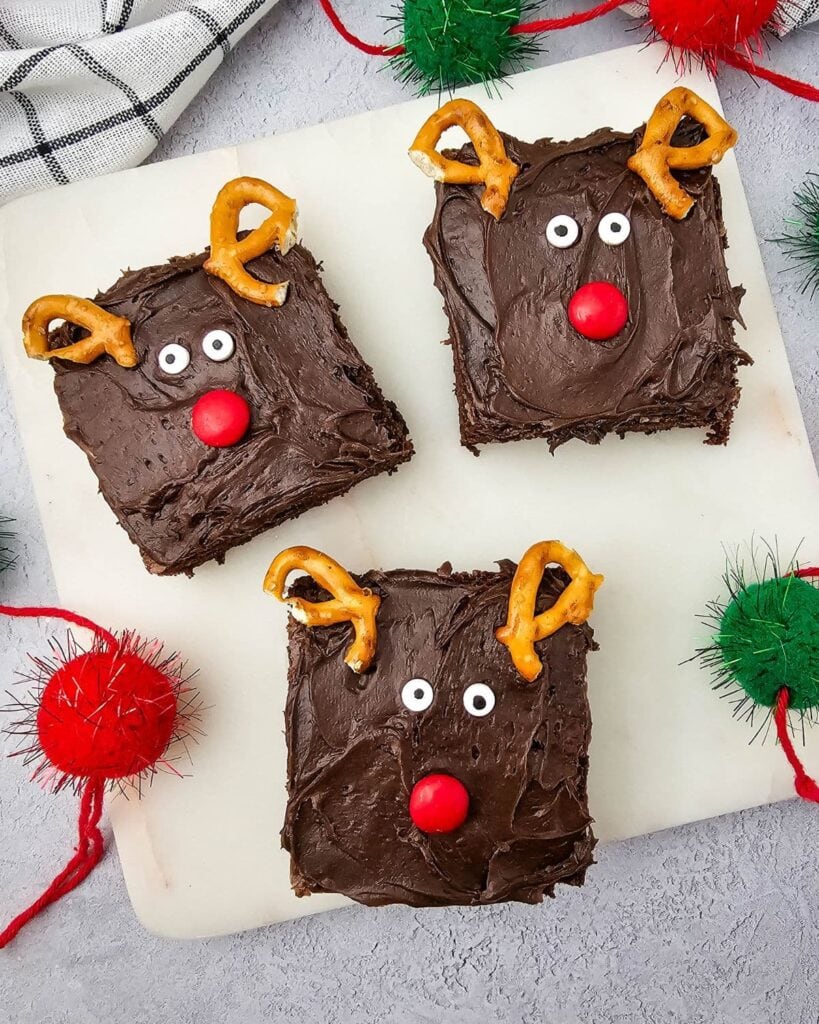 Square chocolate brownies decorated as reindeer with pretzel antlers, candy eyes, and red noses, arranged on white surface with festive pom-poms and checkered cloth.