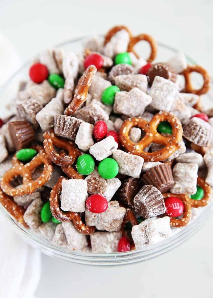 Clear glass bowl of festive snack mix with powdered cereal, pretzels, peanut butter cups, and red and green M&Ms, arranged for Christmas treat presentation.