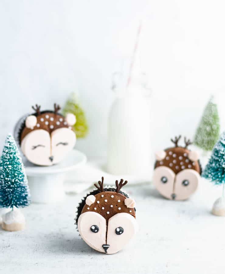 Cupcakes decorated as deer with brown frosting, white snouts, chocolate antlers, and fondant ears, arranged on white surface with miniature trees and milk bottle in festive setting.