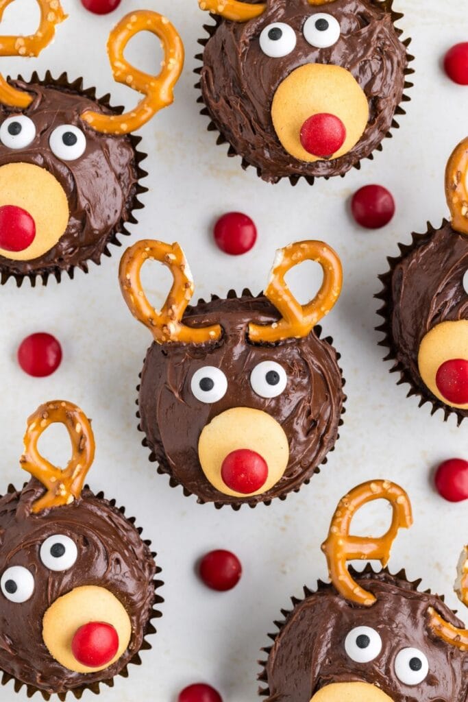 Chocolate cupcakes decorated as reindeer with frosting, candy eyes, wafer snouts, red candy noses, and pretzel antlers, arranged on light surface with scattered red candies.