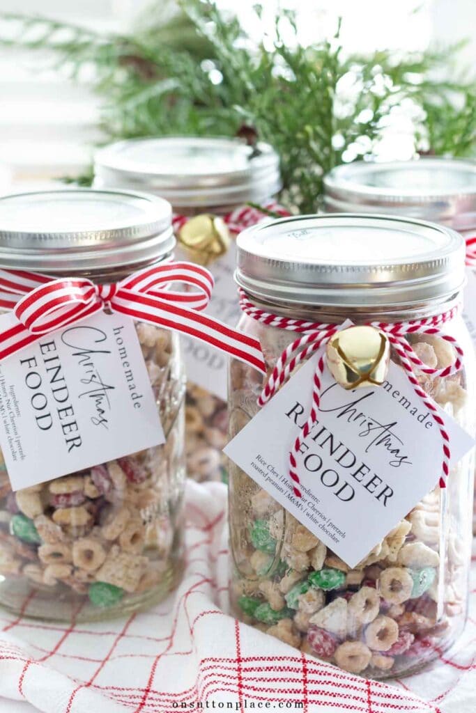 Mason jars of “homemade Christmas Reindeer Food” snack mix with striped ribbons and gold bells, arranged on red grid cloth with greenery in background.