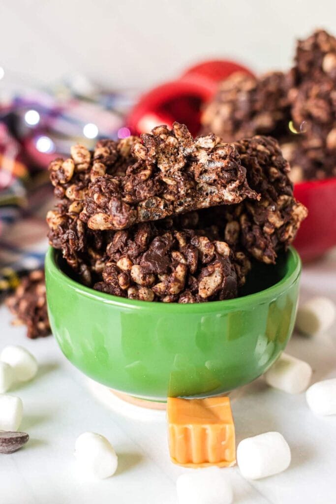 Green mug filled with chocolate-coated rice clusters, surrounded by marshmallows, caramel square, and chocolate chips with blurred holiday décor in background.