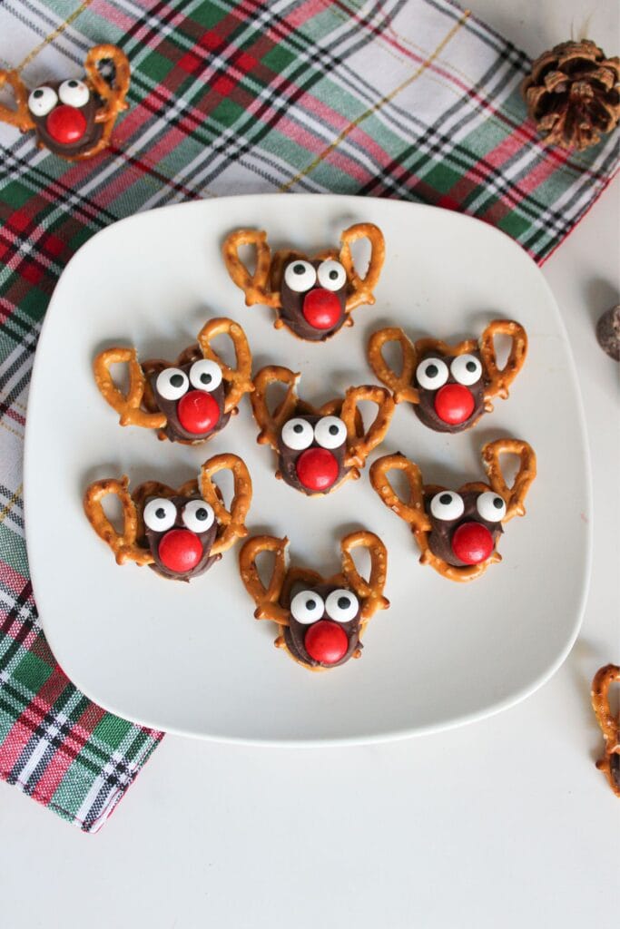 White plate with chocolate reindeer treats made from pretzels, candy eyes, and red noses, set on plaid cloth with pinecone nearby.