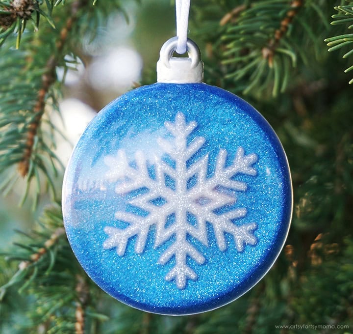 Blue Christmas ornament with glittery white snowflake design hanging on pine branch with green needles.