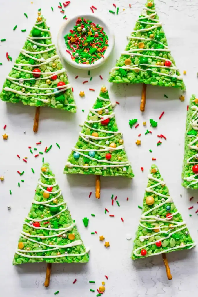 Green Rice Krispies trees decorated with zigzag icing and colorful candy balls, set on pretzel stick trunks, with festive sprinkles scattered on light surface and small bowl of sprinkles nearby.