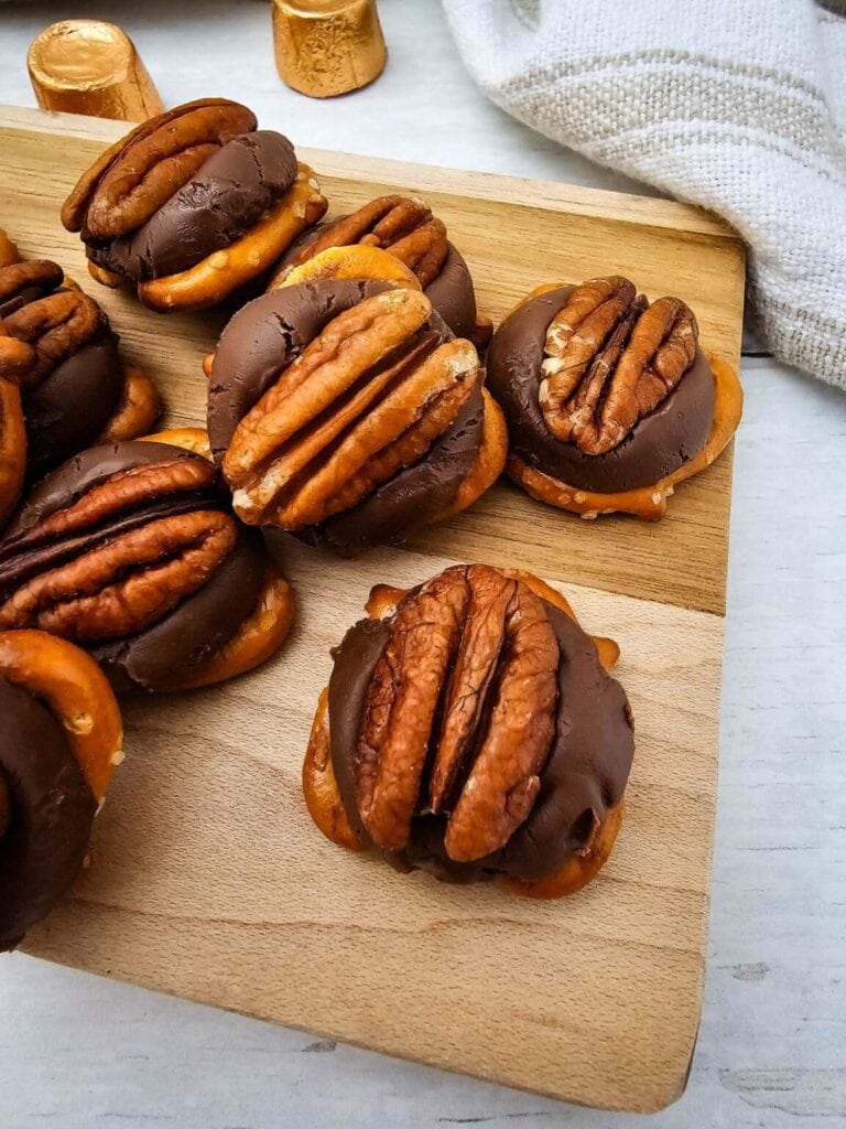 Pretzel-based candies topped with melted chocolate and pecan halves, arranged on wooden board with gold-wrapped caramels and white cloth nearby.