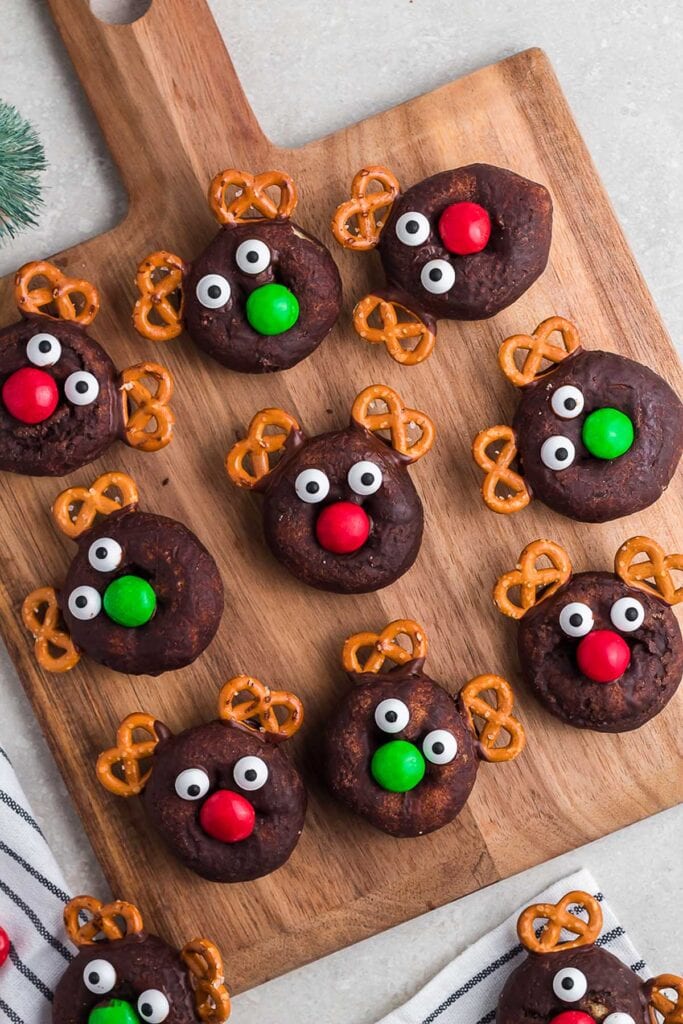 Wooden board of chocolate donuts decorated as reindeer with candy eyes, red or green noses, and pretzel antlers, arranged in festive holiday display.