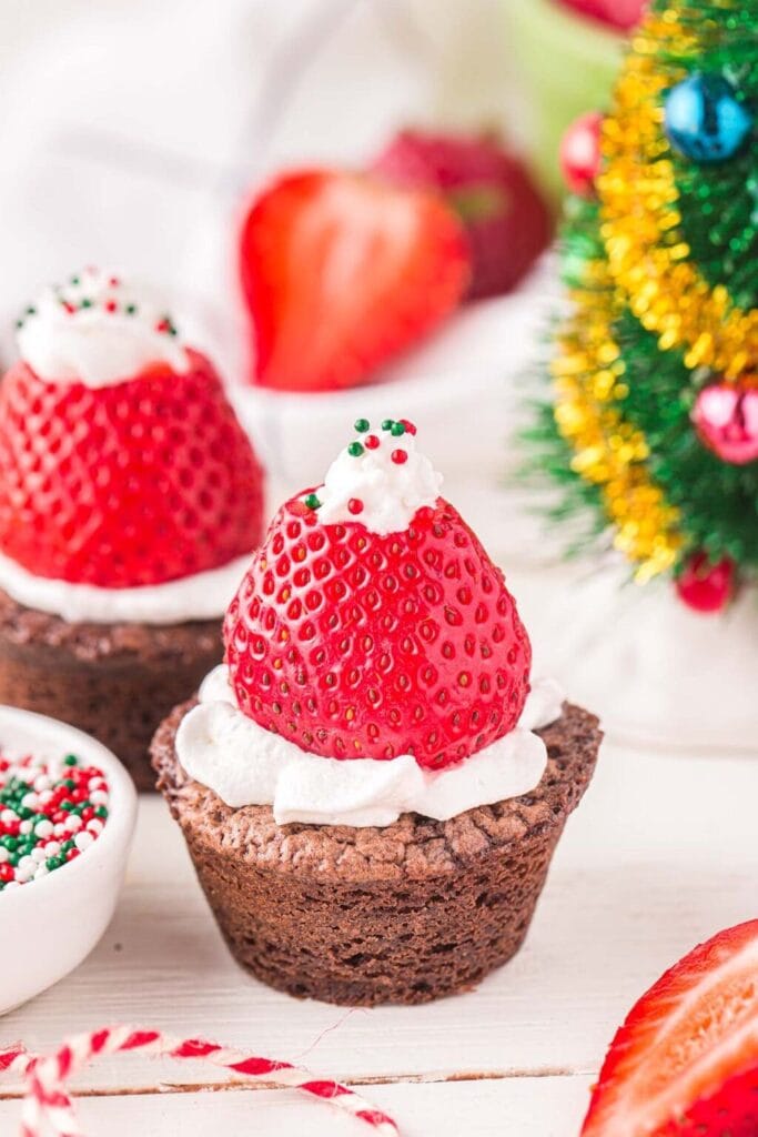 Chocolate brownie cups topped with whipped cream and upside-down strawberries, finished with frosting tip and sprinkles to resemble Santa hats, set beside decorated Christmas tree and bowl of sprinkles.