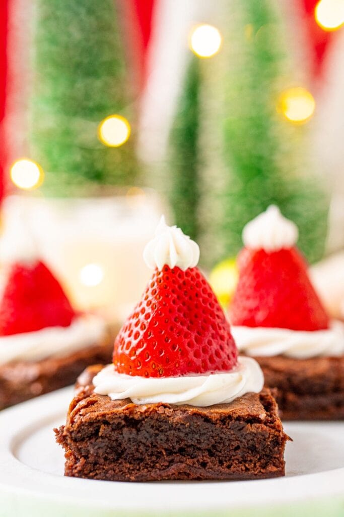 Brownie square topped with white frosting and upside-down strawberry, finished with frosting tip to resemble Santa hat, set against blurred Christmas lights and decorations.