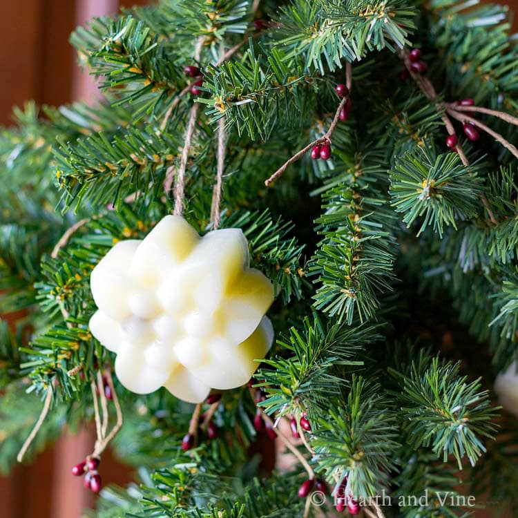 Light yellow flower-shaped ornament made of wax or soap, hanging by twine on pine branch with red berries.
