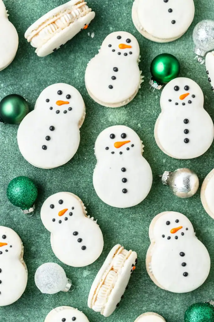 Snowman-shaped cookies with icing faces and buttons, arranged on green surface with festive ornaments scattered around.