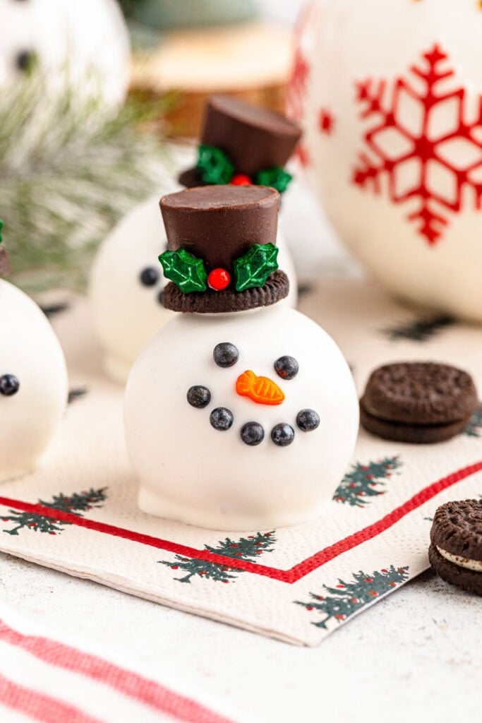 Round white snowman dessert with candy face, carrot nose, and chocolate hat decorated with holly, placed on Christmas napkin with festive background.