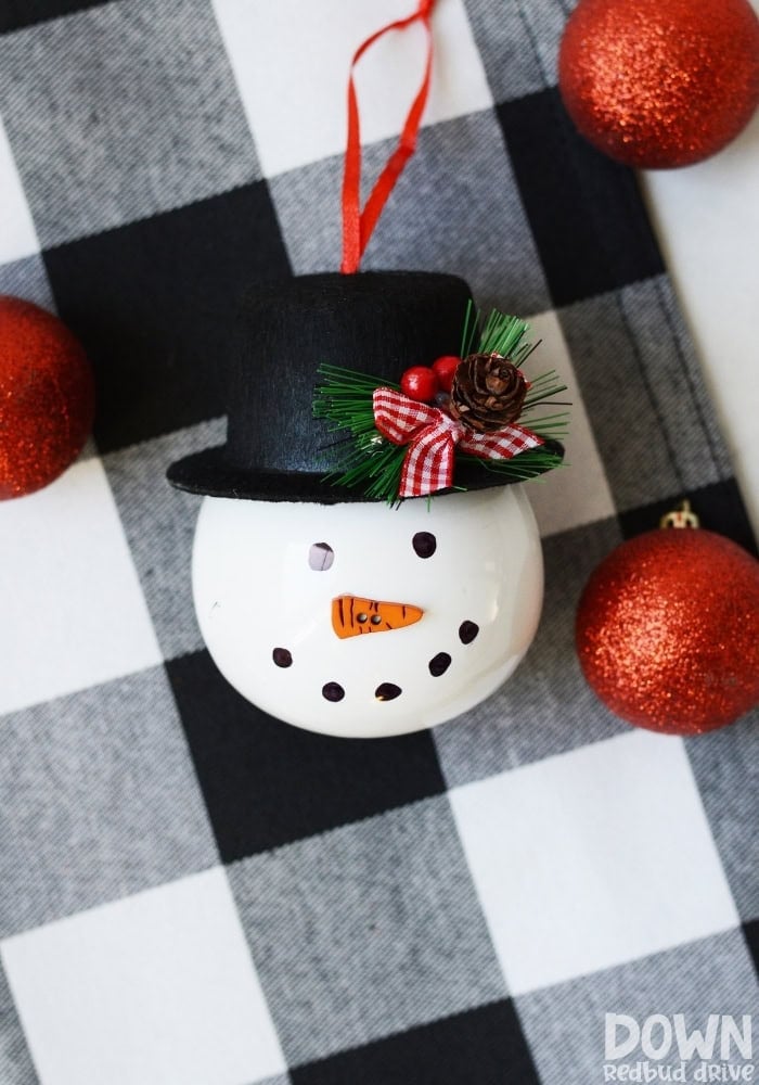 White snowman head ornament with carrot nose, black hat decorated with greenery and berries, and red ribbon loop, placed on checkered fabric with glittery red Christmas balls.