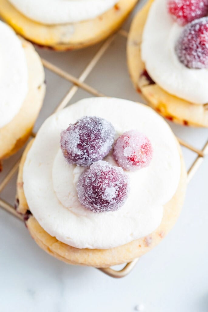 Round frosted cookie topped with sparkling sugared cranberries, resting on cooling rack with other freshly baked cookies nearby.