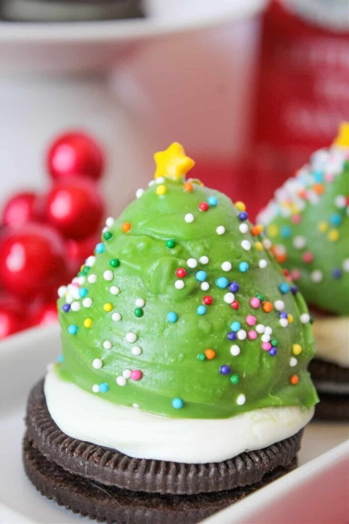 Cookie base topped with green frosting Christmas tree decorated with sprinkles and yellow star, set against festive red ornaments.