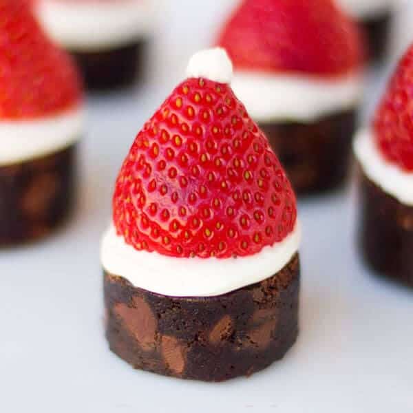 Mini brownie bases topped with white cream and halved strawberries shaped like Santa hats, finished with small cream dollops, arranged in festive holiday setting.