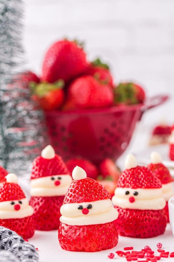 Strawberries decorated as mini Santas with cream beards, hats, candy noses, and eyes, arranged on white plate with festive background.
