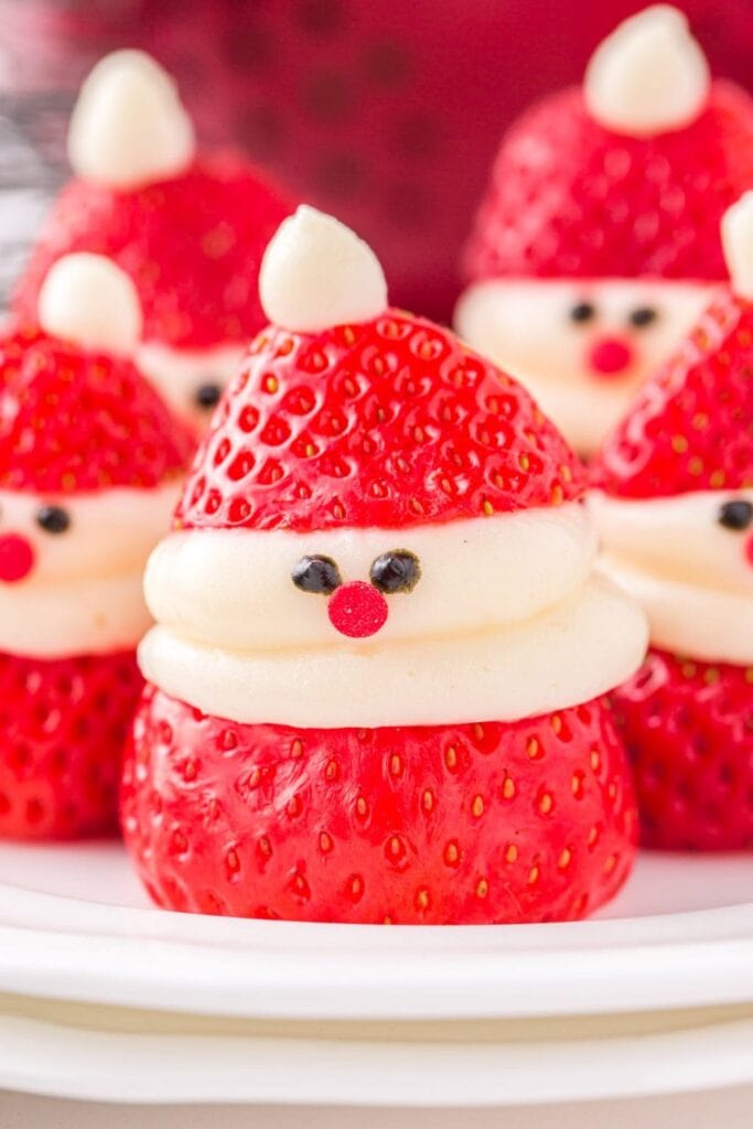 Strawberries decorated as mini Santas with cream beards, hats, candy noses, and eyes, arranged on white plate with festive background.