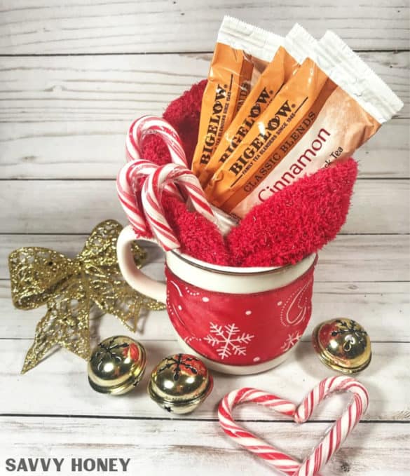 Red snowflake mug filled with tea packets, fuzzy socks, and candy canes, surrounded by gold bow, jingle bells, and heart-shaped candy canes on rustic wood background.