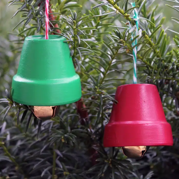 Green and red painted flower pots repurposed as Christmas bell ornaments with golden bells attached, hanging from pine branch.