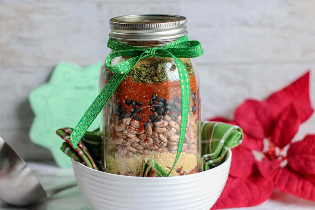 Mason jar layered with colorful beans and lentils, tied with green ribbon, placed in plaid-lined bowl with poinsettia in background.