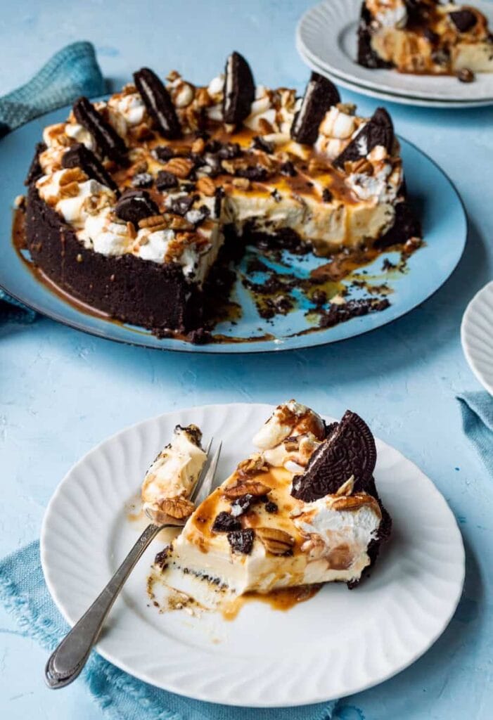 Partially sliced Oreo caramel cheesecake with cookie crust, creamy filling, whipped cream, caramel drizzle, chopped nuts, and Oreo halves, served on blue plate with slices on white plates nearby.