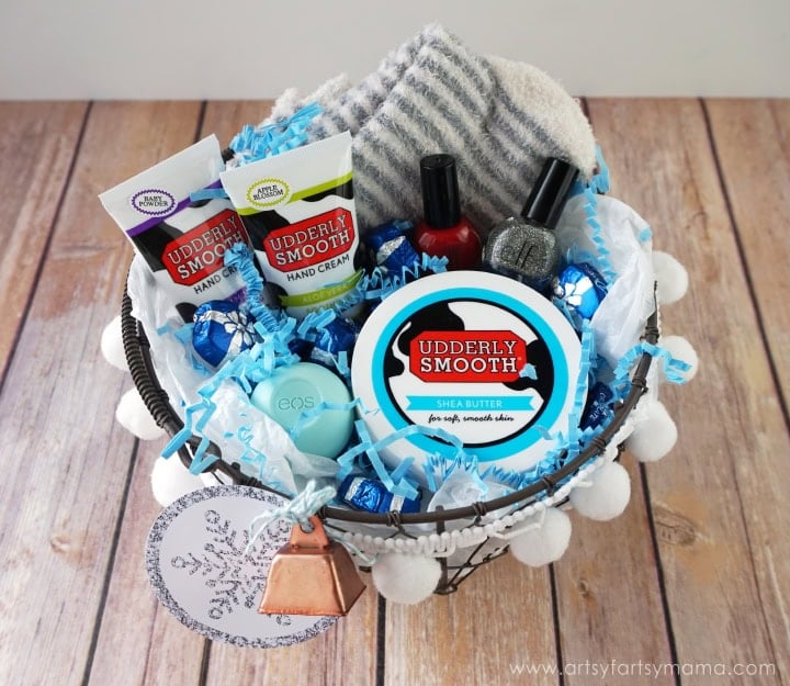 Basket with hand creams, lip balm, nail polish, striped socks, and chocolates, decorated with blue filler, pom-poms, snowflake tag, and bell on wooden surface.