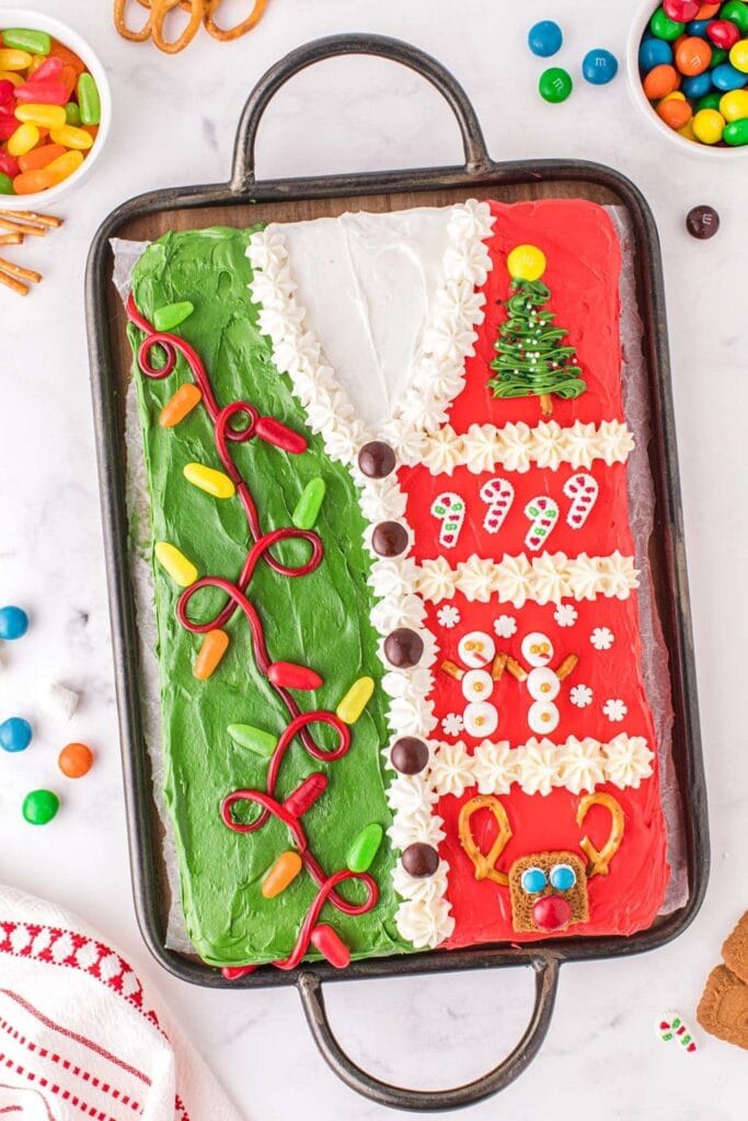 Rectangular cake decorated as colorful Christmas sweater with frosting lights, candy canes, snowmen, reindeer, and tree, set in pan surrounded by bowls of candies.