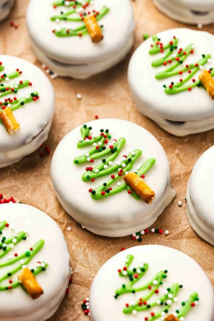 Round sandwich cookies coated in white icing, decorated with green Christmas tree designs made of icing, sprinkles, and pretzel trunks, arranged on parchment paper.