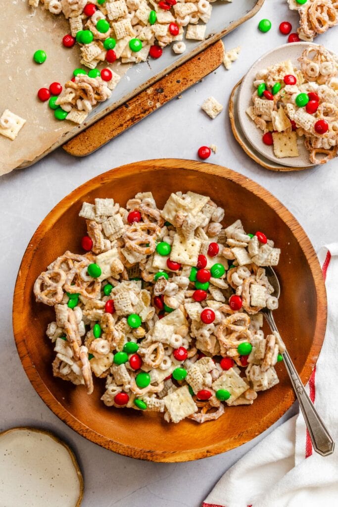 Wooden bowl of holiday snack mix with cereal squares, pretzels, Cheerios, white chocolate-coated pieces, and red and green M&Ms, arranged on baking sheet and plate for festive presentation.