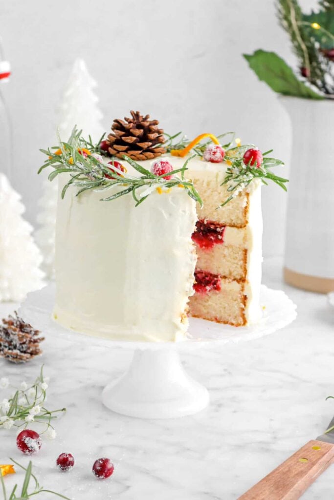 Tall layered cake with white frosting, red berry filling, and festive toppings of sugared cranberries, rosemary, pinecone, and orange peel, displayed on white stand with seasonal decorations.