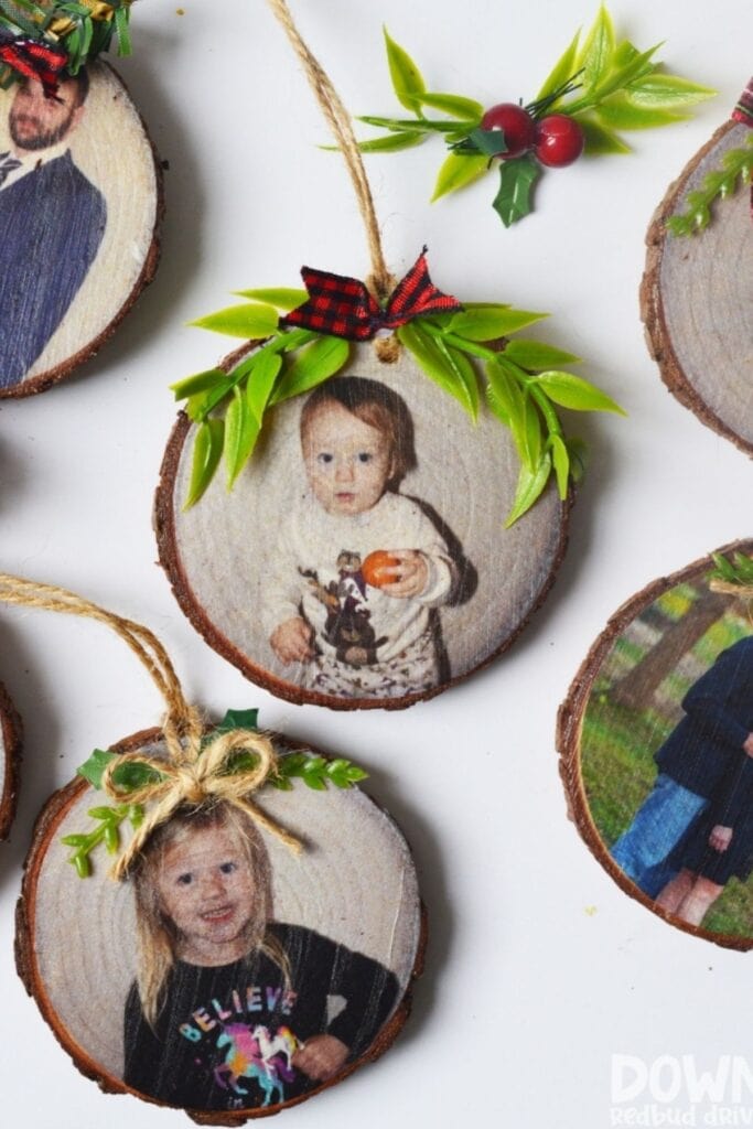 Round wooden ornaments printed with family photos, decorated with greenery and bows, each with twine loop for hanging, arranged on white surface.
