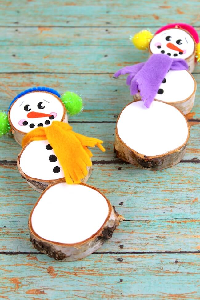 Two handmade snowmen from stacked wood slices with painted faces, colorful felt scarves, and pom-pom hats, placed on turquoise wooden surface.