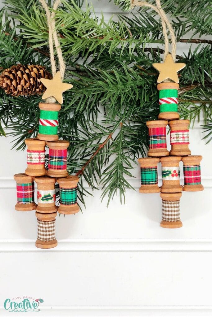 Two handmade Christmas tree ornaments from stacked thread spools wrapped in festive ribbons, topped with golden stars, hanging on pine branch with cone.