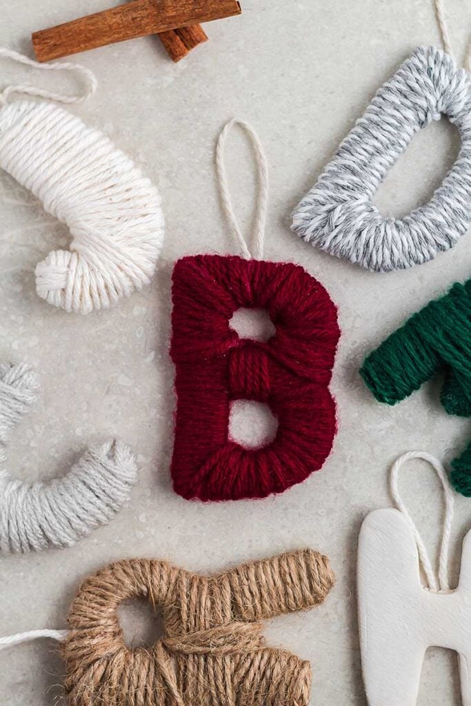 Handmade alphabet ornaments wrapped in colorful yarn with hanging loops, arranged with cinnamon sticks on festive surface.