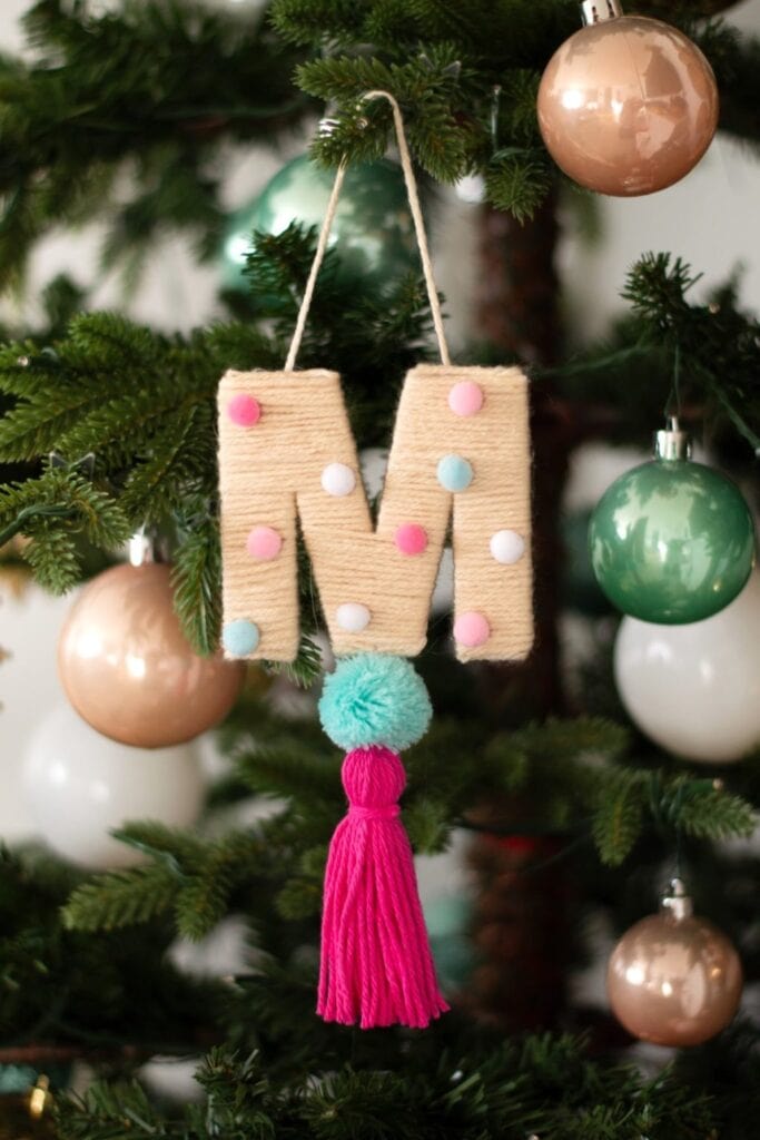 Yarn-wrapped letter “M” ornament with pastel pom-poms, turquoise pom-pom, and pink tassel, hanging on Christmas tree among shiny baubles.