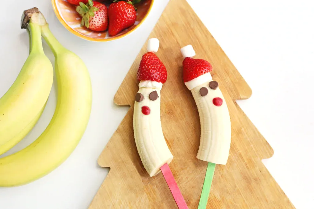 Bananas decorated as Santas with strawberry hats, marshmallow tops, chocolate chip eyes, and red candy noses on Christmas tree-shaped cutting board with strawberries nearby.