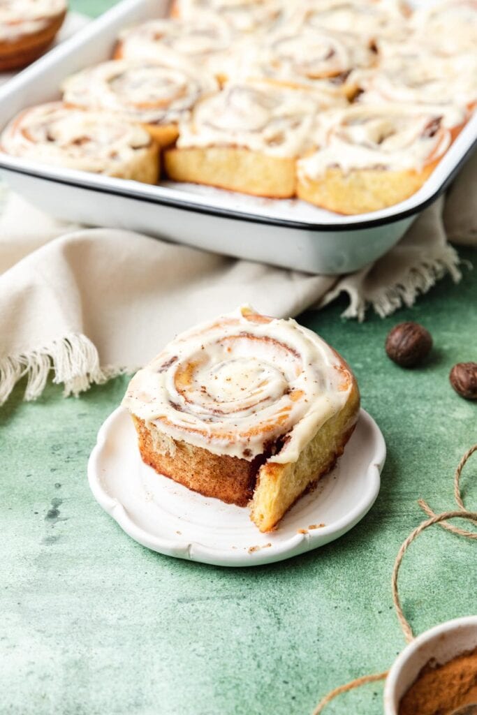 Close-up of cinnamon roll with white icing on plate, baking dish of rolls in background, set on green surface with beige napkin.